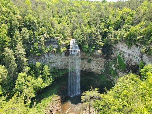 A stunning waterfall cascades into a lush, green forested area, surrounded by rocky cliffs and vibrant foliage.
