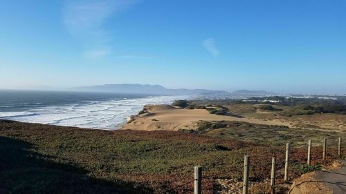 Scenic coastal view with sandy dunes, waves, and distant mountains under a clear blue sky.