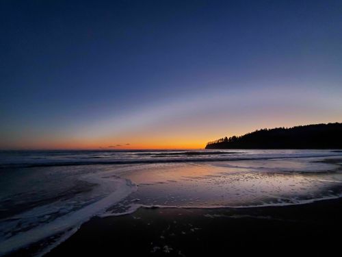 A serene beach at sunset, with gentle waves and a silhouette of trees along the horizon under a colorful sky.