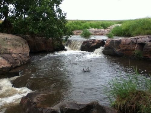 A serene river flows over rocks, surrounded by lush greenery and gentle waterfalls under a cloudy sky.