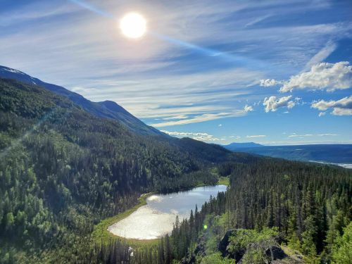 A serene landscape featuring a sunlit lake surrounded by lush forests and mountains under a clear blue sky.