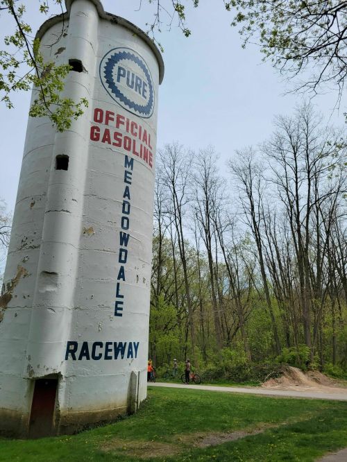 A tall, white gas station silo with "Pure Official Gasoline" and "Meadowdale Raceway" painted on it, surrounded by trees.