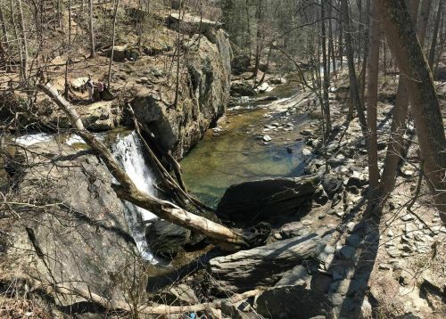 A serene landscape featuring a small waterfall flowing into a clear stream, surrounded by rocky terrain and trees.