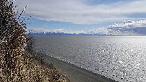 A scenic view of a calm sea with mountains in the distance under a partly cloudy sky. Grass and shoreline in the foreground.