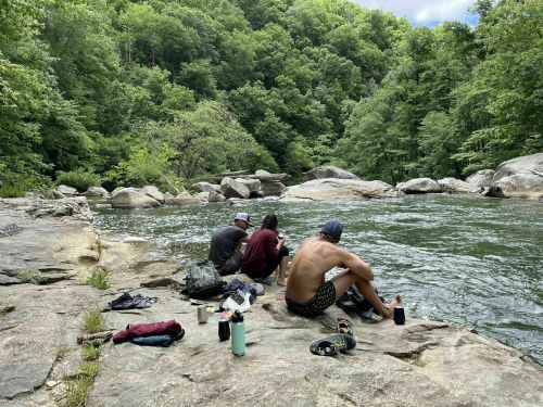 Three people sit on a rocky riverbank, enjoying nature, with lush green trees and water flowing nearby.