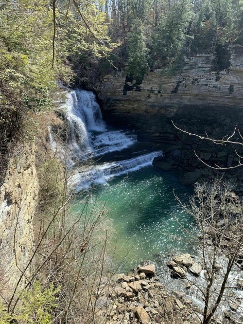 A serene waterfall cascades into a clear pool, surrounded by rocky cliffs and lush greenery.
