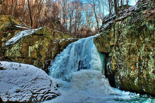 A frozen waterfall cascading over rocky cliffs, surrounded by snow-covered trees and a clear blue sky.