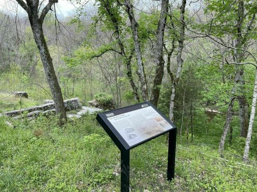 A sign stands in a wooded area, surrounded by trees and greenery, with a stone structure visible in the background.