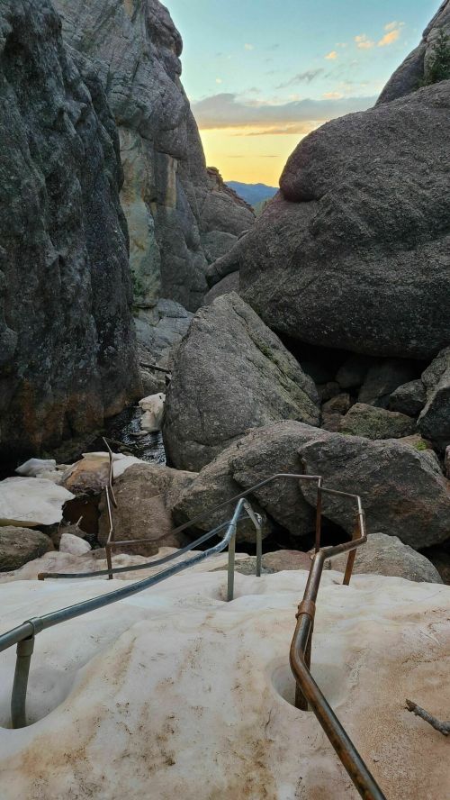 A winding metal staircase leads down through rocky terrain, with large boulders and a sunset in the background.