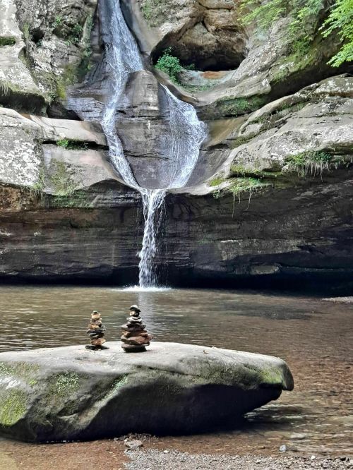 A serene waterfall cascades into a pool, with stacked stones on a rock in the foreground surrounded by lush greenery.