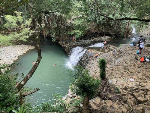 A scenic river with a waterfall, surrounded by lush greenery and people swimming and relaxing along the banks.