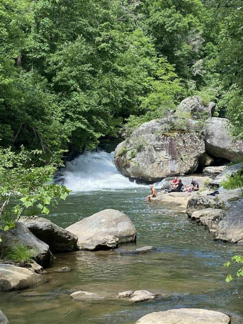 A serene river scene with a waterfall, surrounded by lush greenery and people relaxing on the rocks by the water.