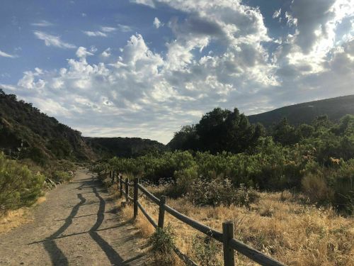 A winding dirt path through lush greenery, framed by mountains under a partly cloudy sky.