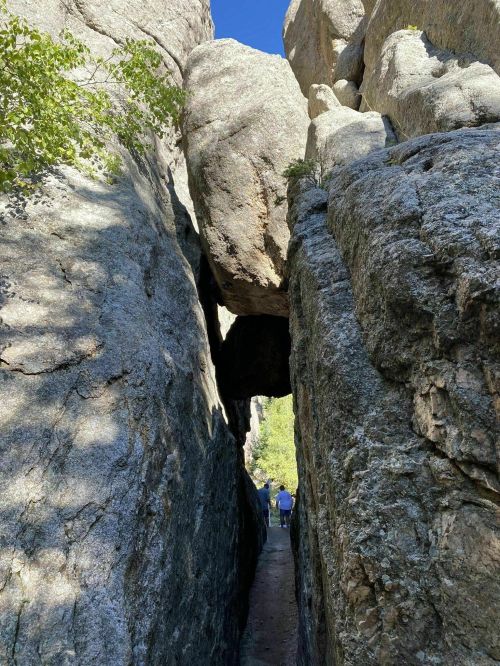 A narrow passage between towering rock formations, with two people walking through under a large boulder overhead.