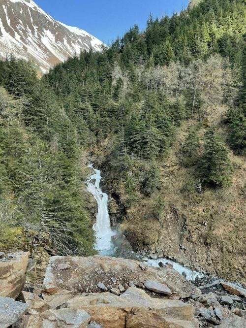 A cascading waterfall flows through a rocky landscape, surrounded by lush green trees and snow-capped mountains.