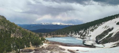 A serene mountain landscape featuring a partially frozen lake surrounded by snow-capped peaks and lush green trees.