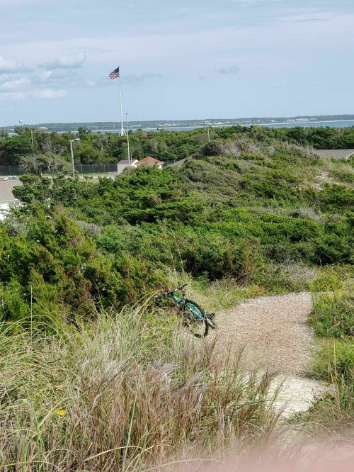 A bike rests on a sandy path surrounded by greenery, with a distant view of a flag and the ocean.