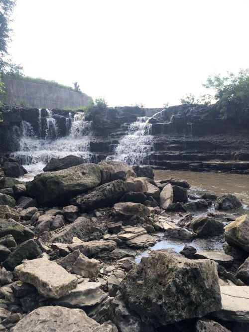 A rocky riverbank with a waterfall cascading over layered stones, surrounded by greenery and a bright sky.