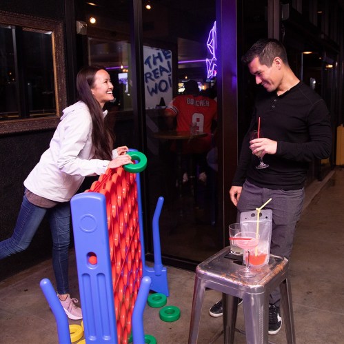 A woman playfully leans on a giant connect-four game while a man smiles at her, holding a cocktail.
