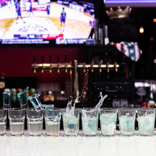A row of shot glasses with colorful liquids, some splashing, in front of a sports TV screen.