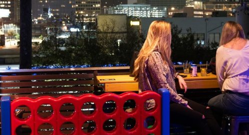 Two people sit at a bar with a city skyline in the background, next to a large Connect Four game.