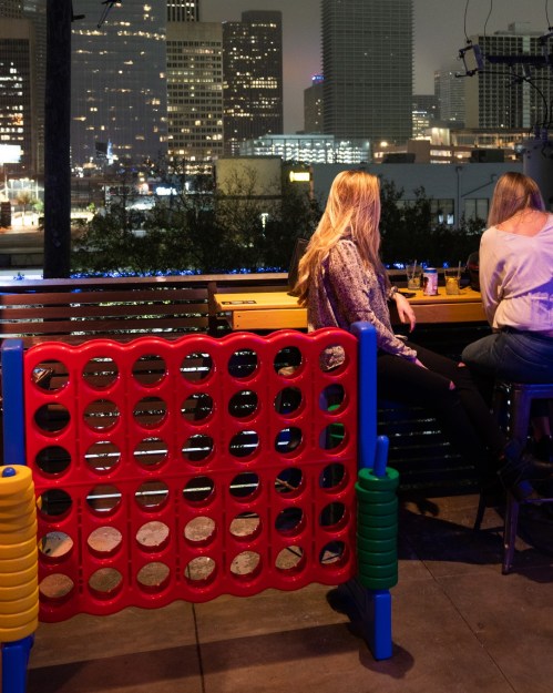 Two women sit at a bar with a city skyline in the background, next to a large Connect Four game.