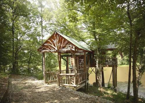 A rustic cabin with a wooden porch, surrounded by trees, overlooking a calm body of water. Sunlight filters through the leaves.
