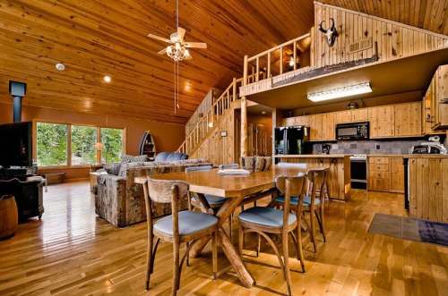 Cozy wooden cabin interior featuring a dining table, kitchen, and loft with a ceiling fan and large windows.