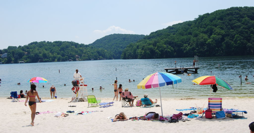 A sunny beach scene with people swimming, lounging under colorful umbrellas, and a lifeguard on duty by the water.