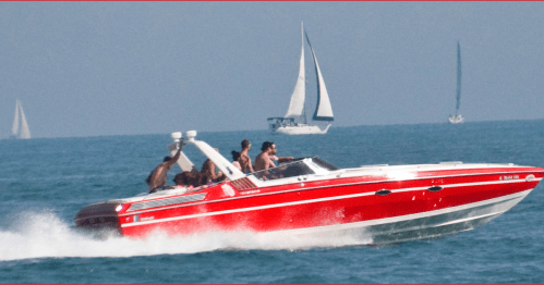A red speedboat with people onboard speeds through the water, with sailboats in the background.
