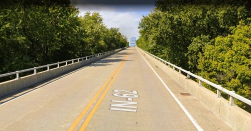 A wide, empty road stretches ahead, flanked by trees, leading to a bridge in the distance.
