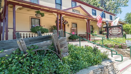 Front view of a charming hotel with a porch, flags, and lush greenery surrounding the entrance.