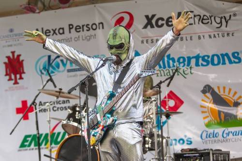 A performer in a silver alien costume plays guitar on stage, with arms raised, in front of a band and sponsor banners.