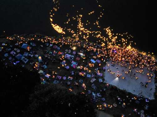 Aerial view of a lakeside gathering with glowing lanterns and blankets spread out on the ground at dusk.