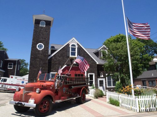 A vintage red fire truck with flags parked in front of a wooden fire station on a sunny day.