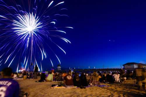 Crowd gathered on a beach watching colorful fireworks illuminate the night sky over a pier.