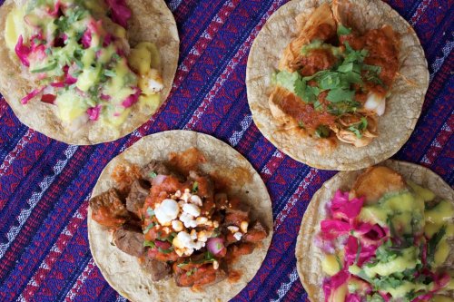 Four colorful tacos on a vibrant woven fabric, topped with various ingredients like meat, salsa, and herbs.