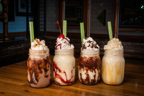 Four colorful milkshakes in mason jars, topped with whipped cream, syrups, and garnishes, on a wooden table.