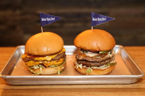 Two gourmet burgers on a metal tray, each topped with a flag, featuring layers of beef, cheese, and fresh toppings.