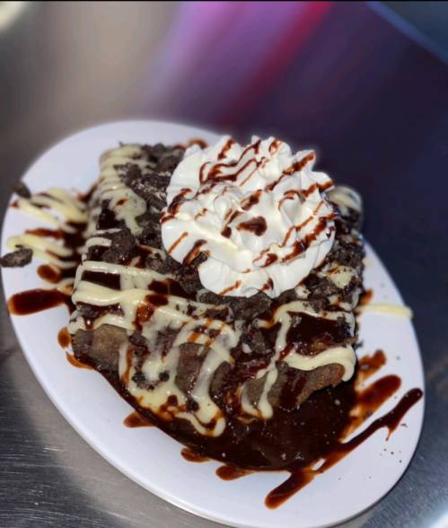 A dessert plate featuring a chocolate brownie topped with whipped cream, chocolate drizzle, and crushed cookies.