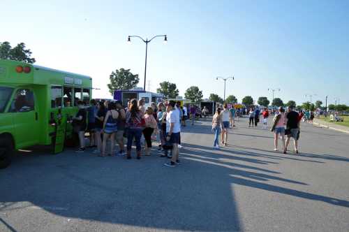 A crowd gathers around food trucks at an outdoor event on a sunny day, with trees and streetlights in the background.