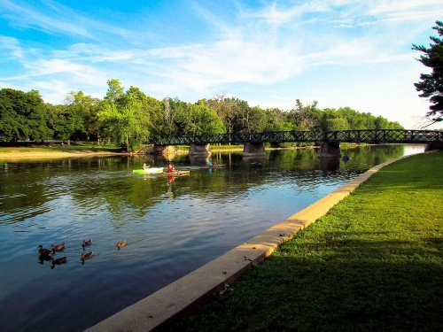 A serene river scene with a bridge, a kayaker, and ducks swimming near the grassy bank under a blue sky.