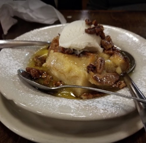 A dessert featuring a warm bread pudding topped with vanilla ice cream and chopped pecans, served on a plate.