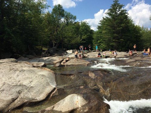 People enjoying a sunny day by a rocky river, surrounded by trees and blue skies.