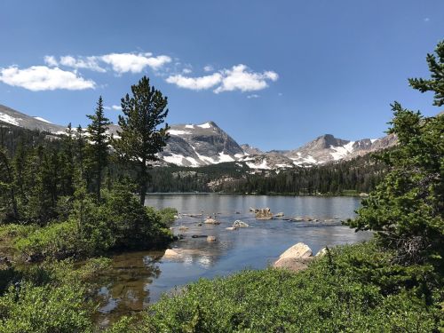 A serene mountain lake surrounded by trees and rocky shores, with snow-capped peaks under a clear blue sky.