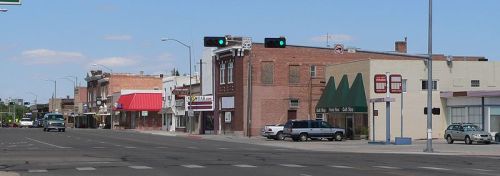 A quiet street scene featuring small businesses, traffic lights, and parked cars under a clear blue sky.