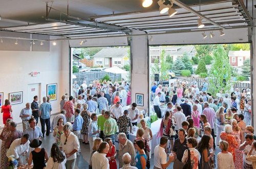 A large crowd gathers in an art gallery, enjoying an event with food, drinks, and colorful artwork on display.