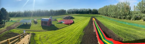 A panoramic view of a green field featuring a colorful waterslide, a play structure, and a small barn.