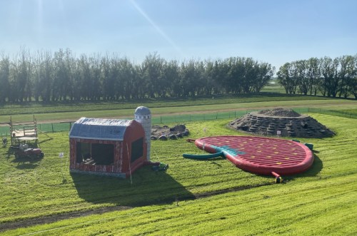 A colorful inflatable slide shaped like a watermelon next to a barn and a dirt mound on a grassy field.