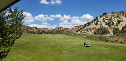 A scenic golf course with rolling hills, blue skies, and a golf cart on the green. Fluffy clouds float above.